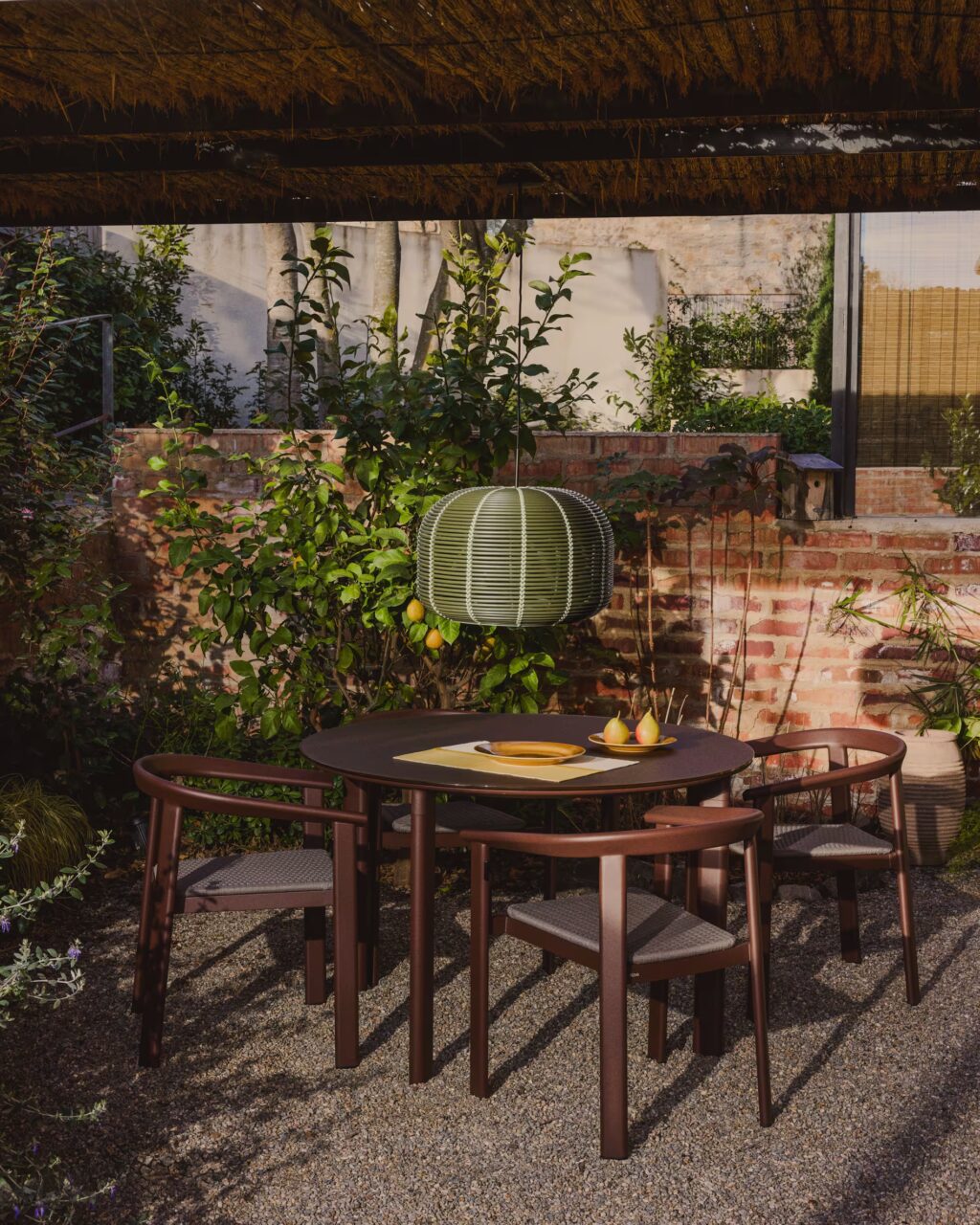 Comedor exterior rústico con mesa redonda y sillas de aluminio efecto madera en jardín con pared de ladrillo.