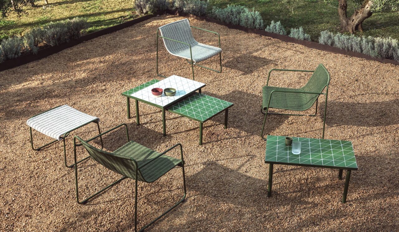 Conjunto de sillas metálicas y mesas bajas con azulejo verde sobre superficie de grava, delimitado por bordes de acero y vegetación estructurada en jardín de diseño contemporáneo.