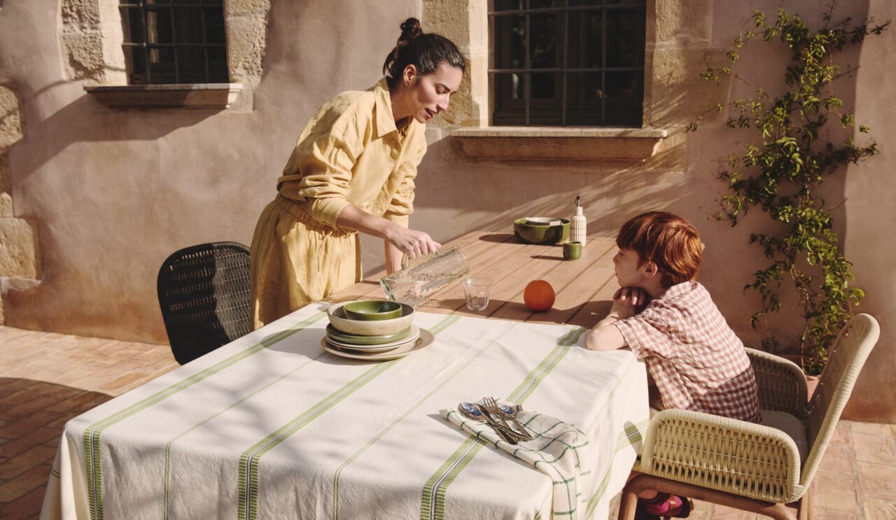 Mujer sirviendo agua a un niño en una mesa de exterior con mantel blanco de rayas verdes en patio soleado.